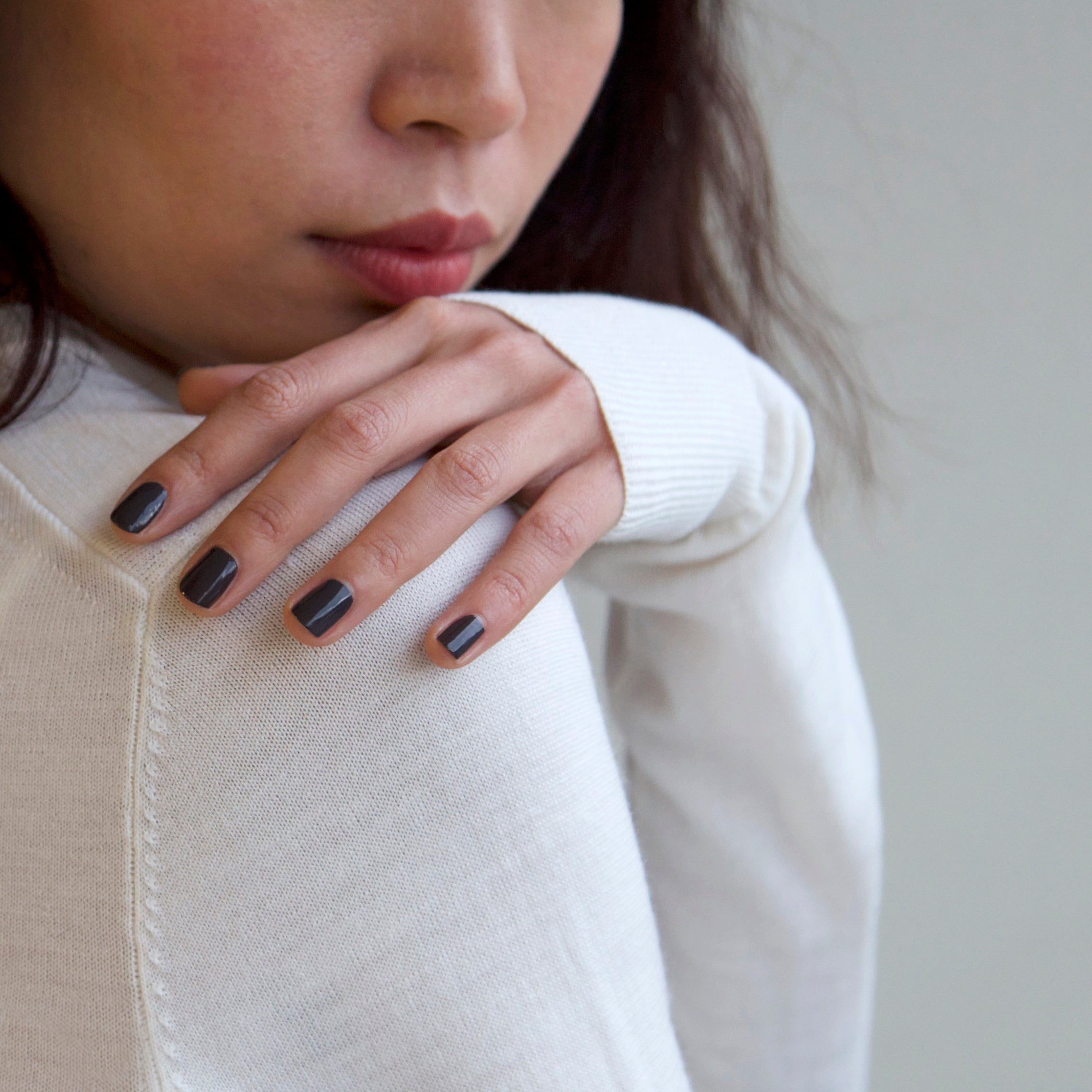 A female resting her hand on her shoulder wearing Volcanic Ash, a dark grey nail polish. Contrasting with her white top and grey background.