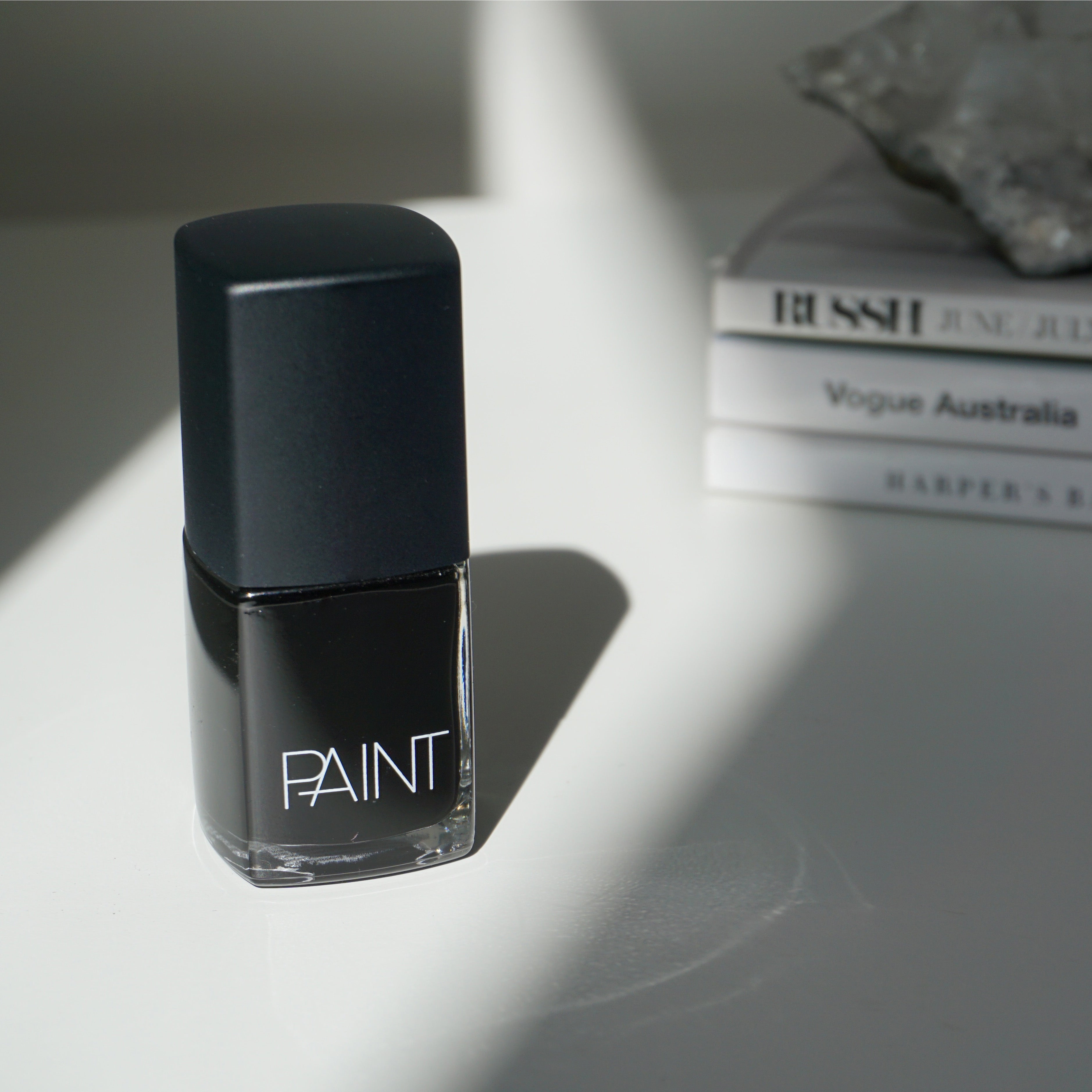 A bottle of Black Dog nail polish by Paint Nail Lacquer against a white backdrop with the sunsetting on the polish, this shade is a classic black colour  