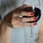 A female hand holding a glass of red wine, wearing Paint Red nail polish, a classic bold red shade