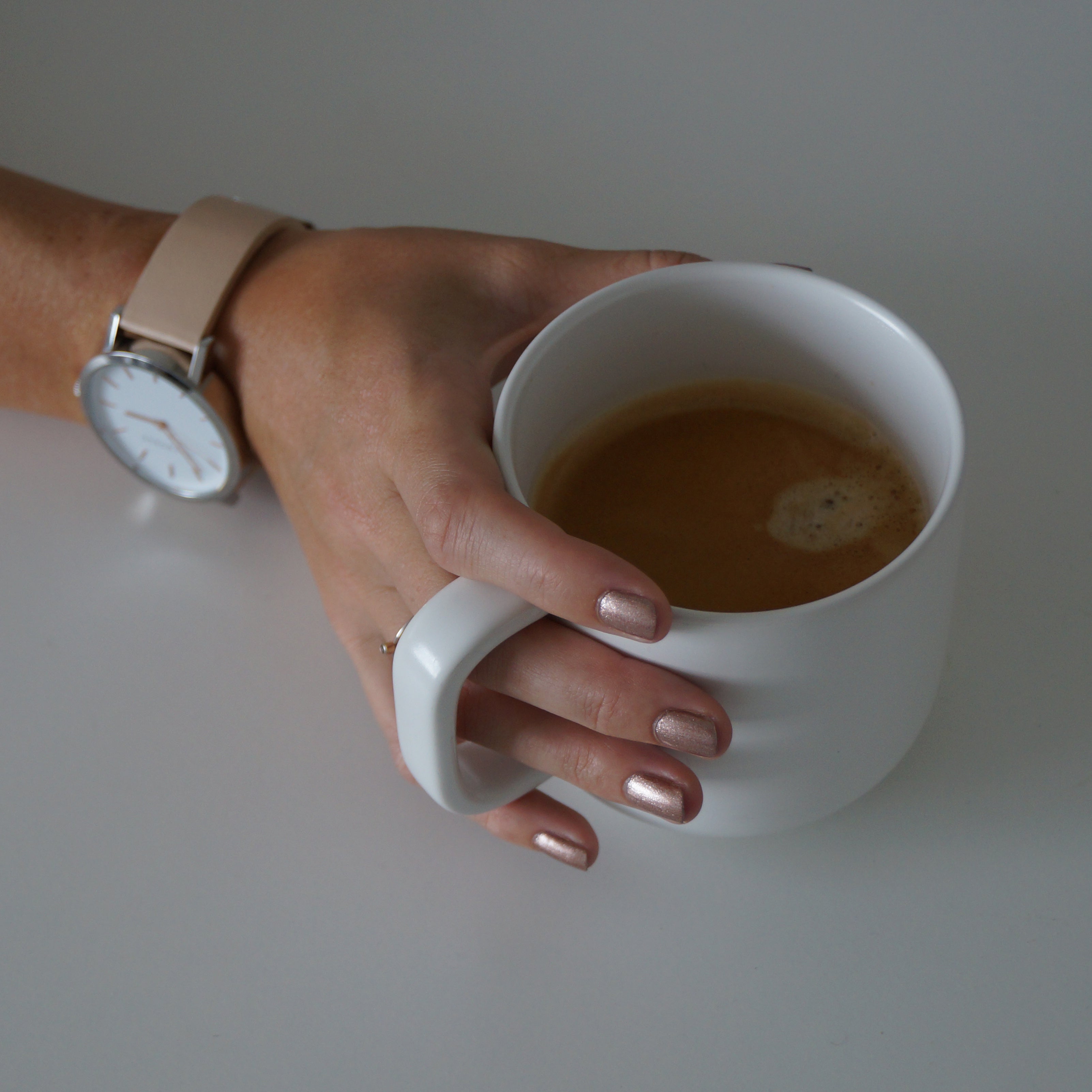 A female hand holding a cup of coffee, wearing Copper Blush nail poilsh - a metallic dark pink colour with gold undertones