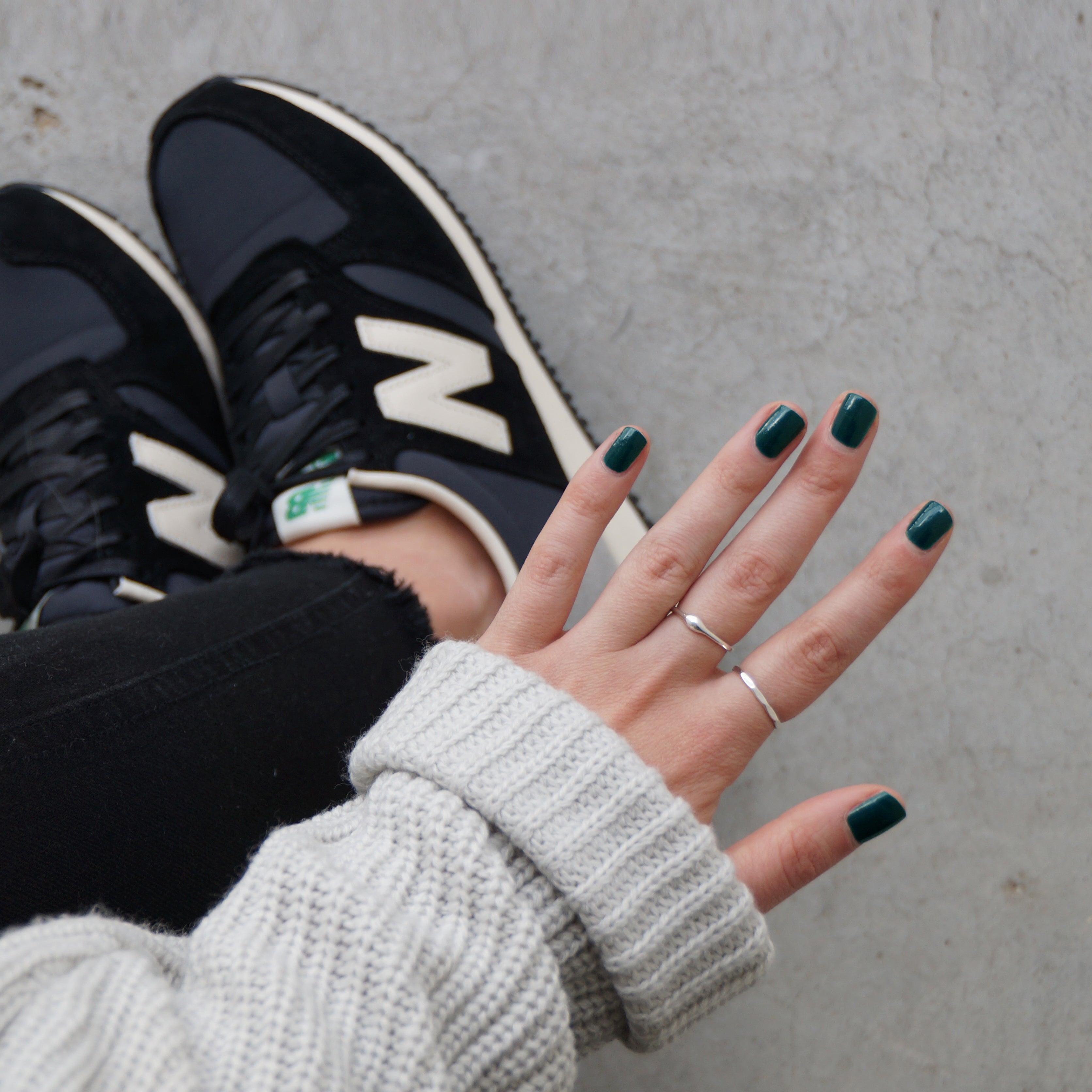 A female holding out her hand, wearing Sea Queen, a dark green nail polish. Contrasting against her black jeansm New Balance shoes, and light grey concrete in the background.