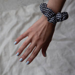 A female hand streched out against a grey backdrop, wearing Pale Glacier a baby blue nail polish and a black and white gingham scrunchie on her wrist