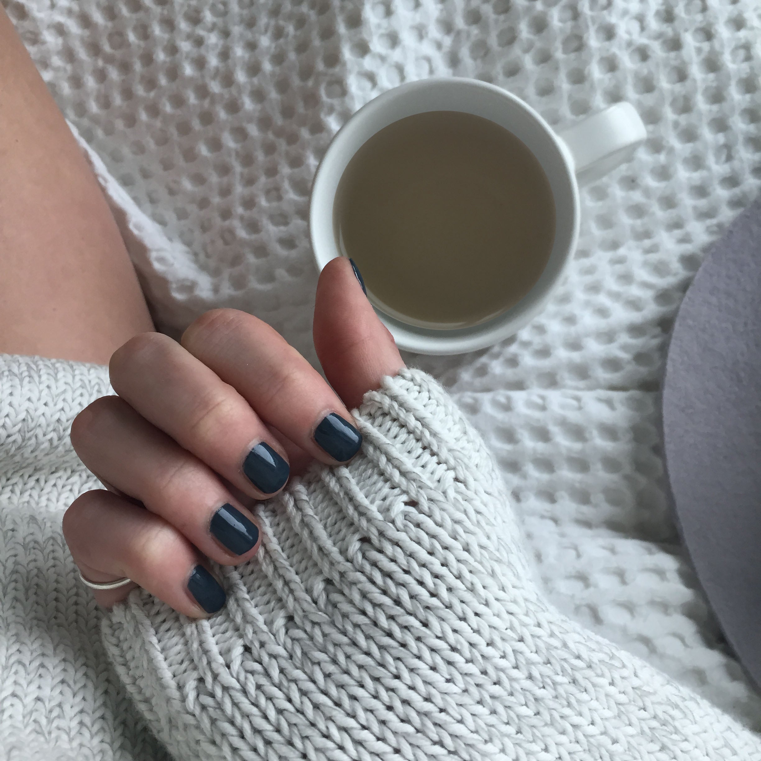 A female clutching the sleeve of her white woolen sweater wearing Storm Grey, a dark blue grey nail polish with green undertones. Constrasting to her cup of tea and blanket in the background.