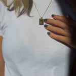 A female with her hand on her chest near her gold necklaces wearing Amazon Haze a dark grey nail polish with green undertones