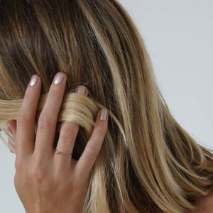 A female side on view holding a lock of hair whilst wearing Copper Blush nail polish, a metallic dark pink shade with gold undertones