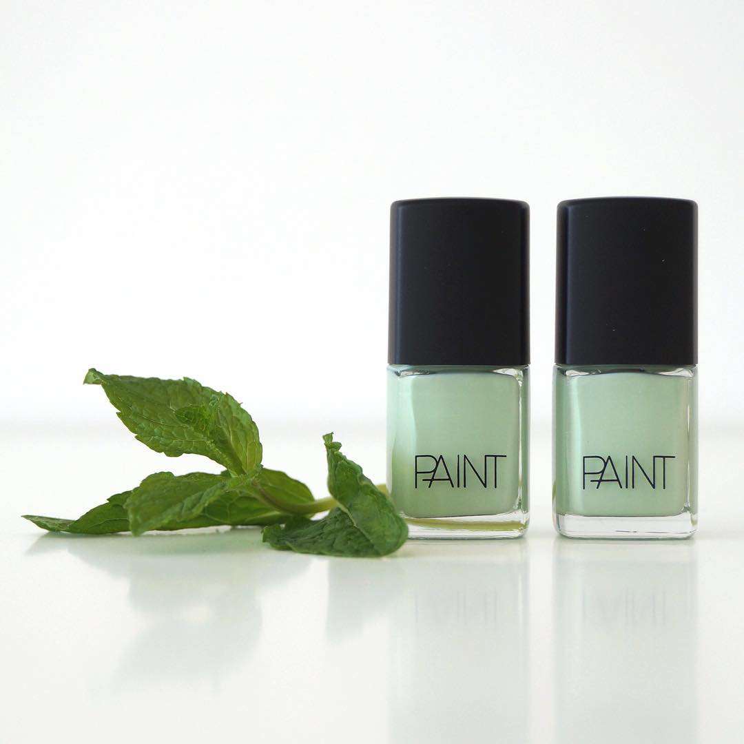 Two bottles of Mint Sorbet Nail Polish by Paint Nail Lacquer against a white backdrop, this shade is a light emerald leafy pastel green in colour. The bottles are next to fresh mint leaves