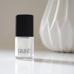 A bottle of Limed White Nail Polish by Paint Nail Lacquer positioned on a white benchtop. This shade is a bold, pearly white colour. In the background is a white detailed vase.