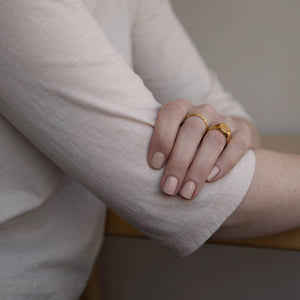 A female with her arms crossed in front, wearing two gold rings and Paint Naked nail polish a light nude colour