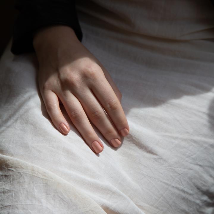 A female hand resting on linen in the sunlight wearing Peach Club nail polish - a light orange cream colour with pink undertones