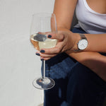 A female holding a glass of white wine in the sunlight, wearing Storm Grey nail polish, a dark blue grey shade.