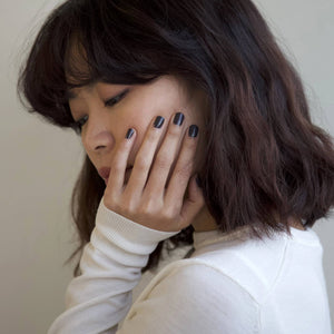 A female holding her face in her hand, wearing Volcanic Ash nail polish a dark grey shade with purple tones